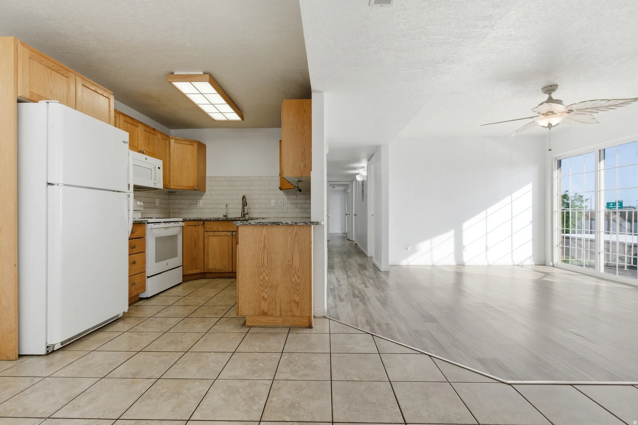 Combined kitchen and living area featuring light wood-finish cabinetry, granite-style countertops, and white appliances