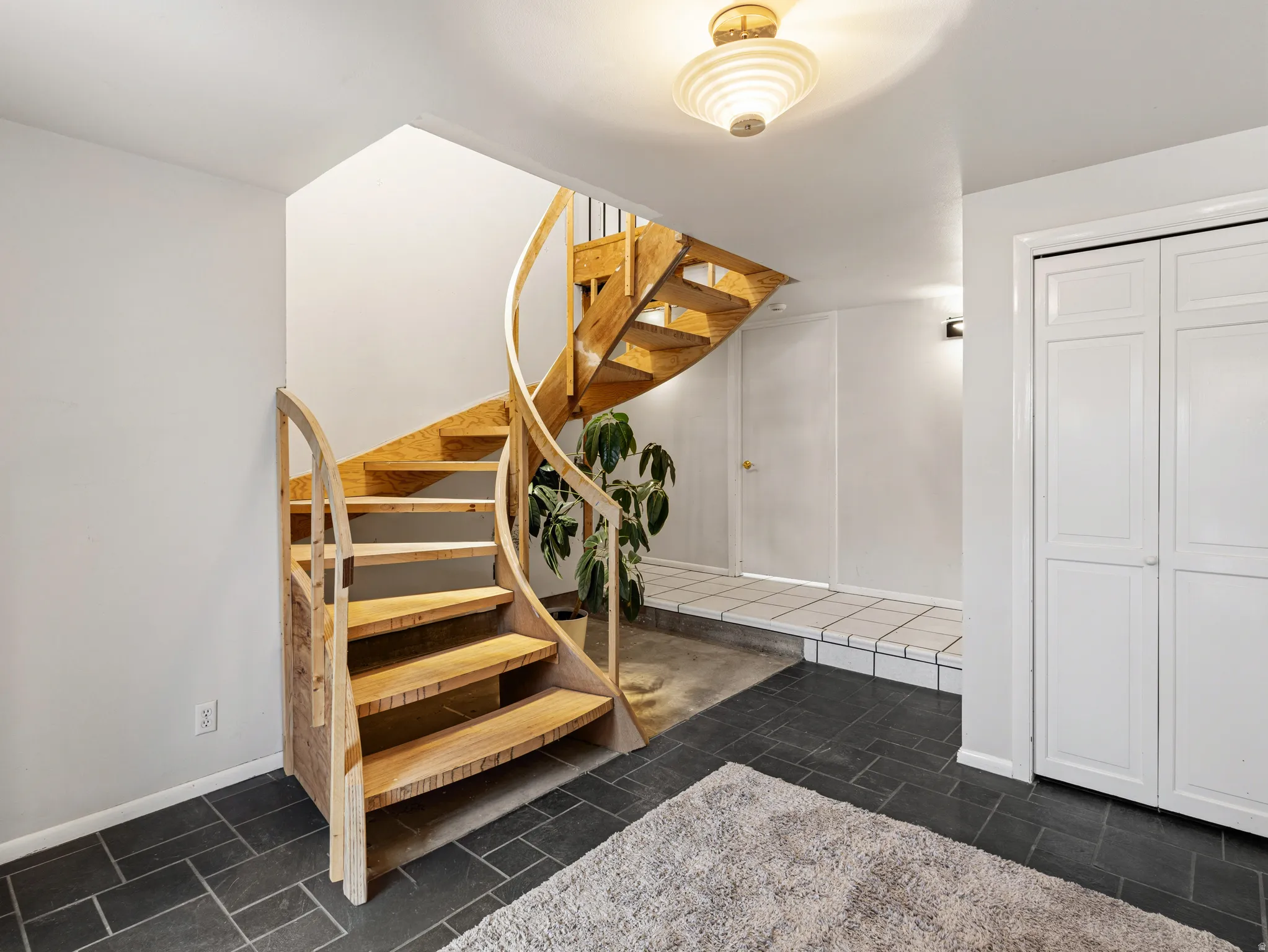 Spiral staircase with natural wood treads and balusters, dark slate-style floor tiling, white walls, built-in bi-fold closet doors, and an overhead semi-flush mount light fixture