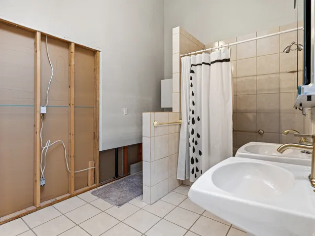 Two wall-mounted pedestal sinks with brass-finish faucets, a tiled shower area, and light-toned floor tiling