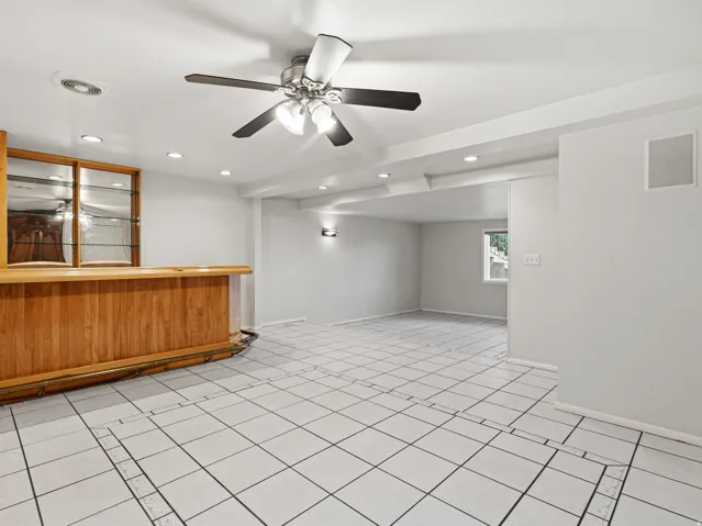 Spacious open room featuring a built-in wood bar with mirrored shelving, white tiled flooring, recessed lighting, and a ceiling fan