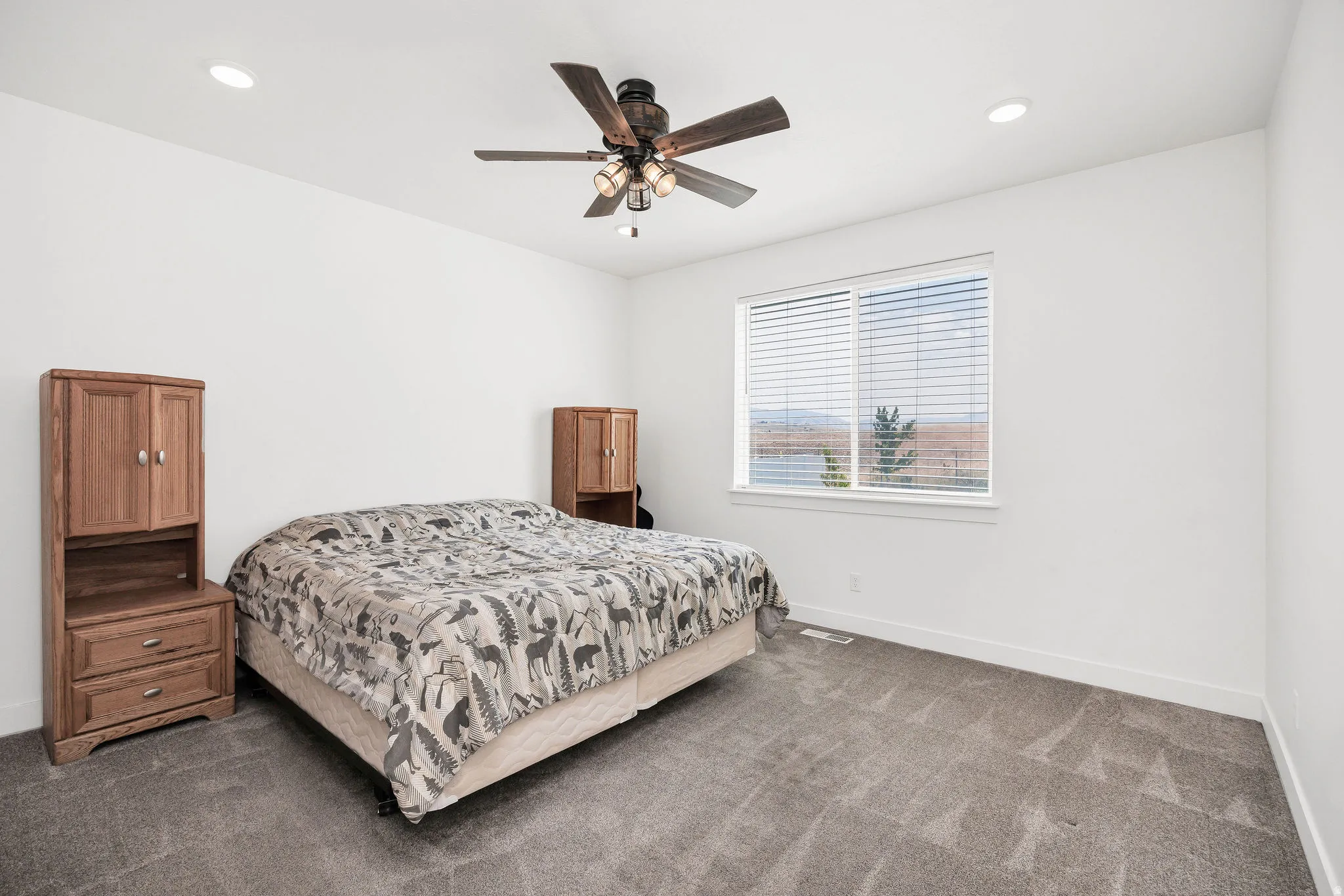 Spacious bedroom featuring plush gray carpeting, a large window with horizontal blinds, and a ceiling fan with integrated lighting