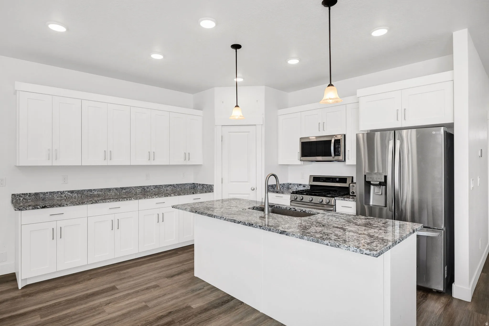 Spacious kitchen featuring white shaker-style cabinetry, patterned countertops, a central island with an undermount sink, stainless steel appliances, and wood-finish flooring