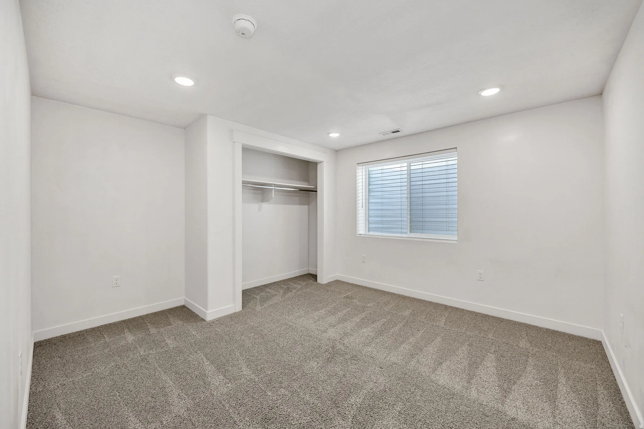 Neutral-toned carpet flooring featuring white baseboards, a built-in closet with shelving, a window with horizontal blinds, and recessed ceiling lighting