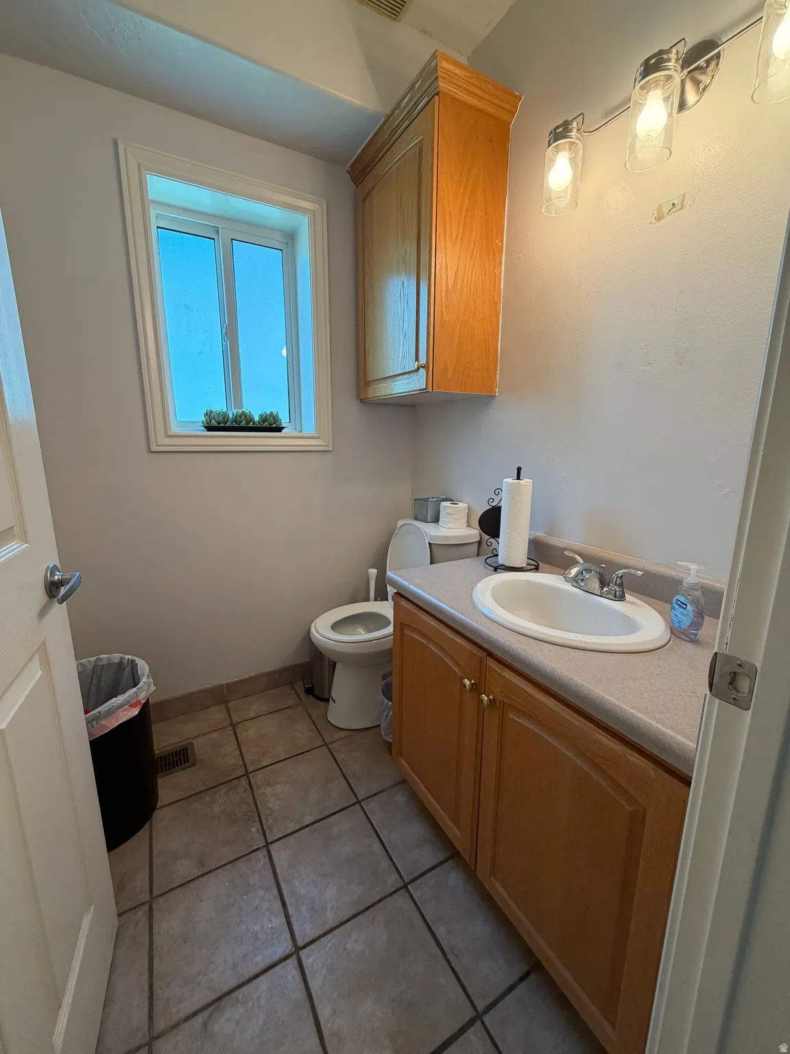 Bathroom featuring a wood-finish vanity with an integrated sink, a matching wood-finish wall cabinet, a window with white trim, a toilet, and neutral-toned tile flooring