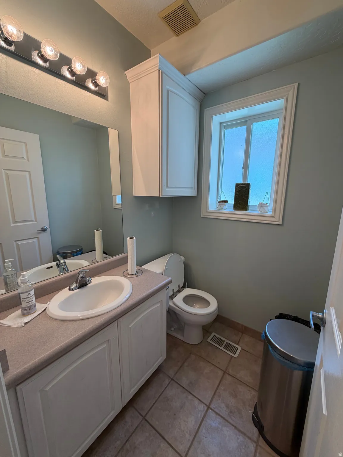 Bathroom featuring a single vanity with an integrated sink, a rectangular mirror, overhead vanity lighting, a window with white trim, and tile flooring