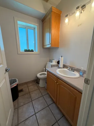 Bathroom featuring a wood-finish vanity with an integrated sink, a matching wood-finish wall cabinet, a window with white trim, a toilet, and neutral-toned tile flooring
