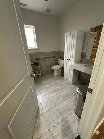 Bathroom with wood-finish tile flooring, a window, a white vanity with an integrated sink, a toilet, and a tall white storage cabinet