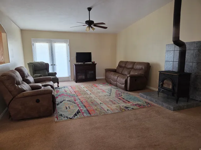 Spacious living area featuring a vaulted ceiling with a ceiling fan, a prominent wood-burning stove with a tiled hearth and wall surround, and double French doors providing outdoor access