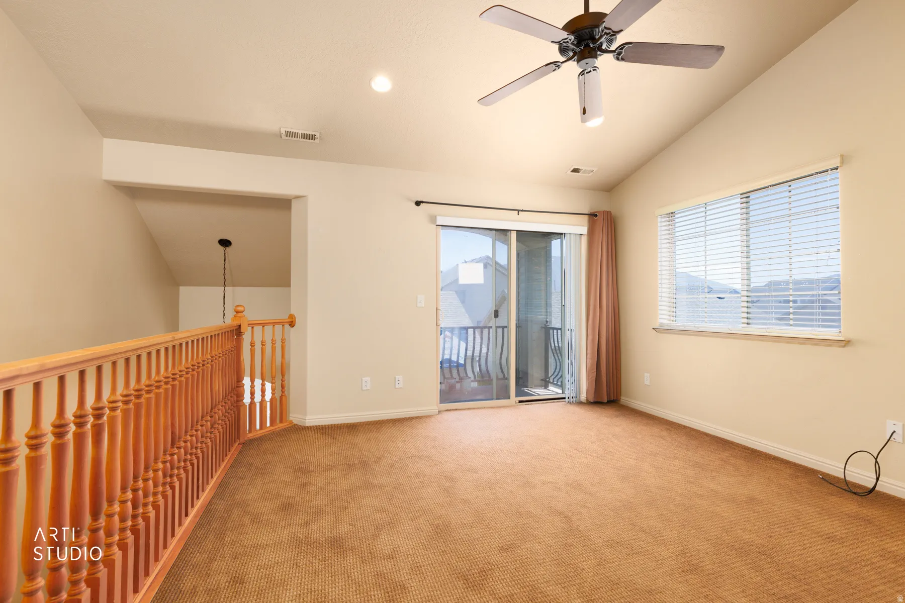 Spacious carpeted room featuring a vaulted ceiling with a ceiling fan, a sliding glass door to a balcony, a large window with blinds, and a wood-finish railing overlooking a lower level