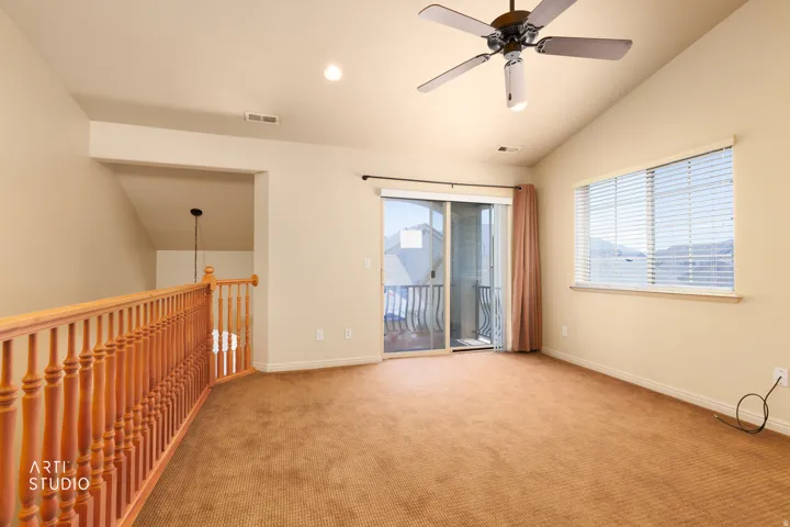 Spacious carpeted room featuring a vaulted ceiling with a ceiling fan, a sliding glass door to a balcony, a large window with blinds, and a wood-finish railing overlooking a lower level