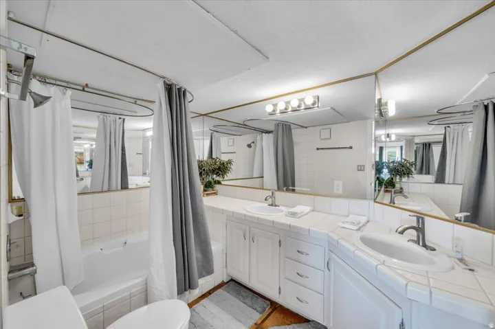 Bathroom featuring a built-in tub with white tile surround, a dual vanity with white tile countertops and integrated sinks, extensive wall-mounted mirrors, and white cabinetry with brushed nickel hardware