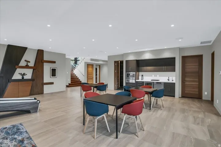 Spacious open area featuring wood-finish flooring, a contemporary wet bar with integrated appliances, recessed lighting, and a prominent wooden staircase