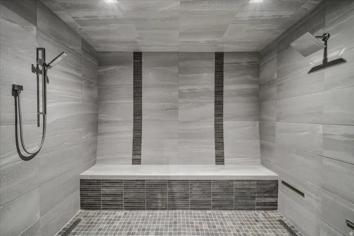 Spacious shower featuring full-height stone-look tile, a built-in bench, dual showerheads with a handheld sprayer, and mosaic tile accents