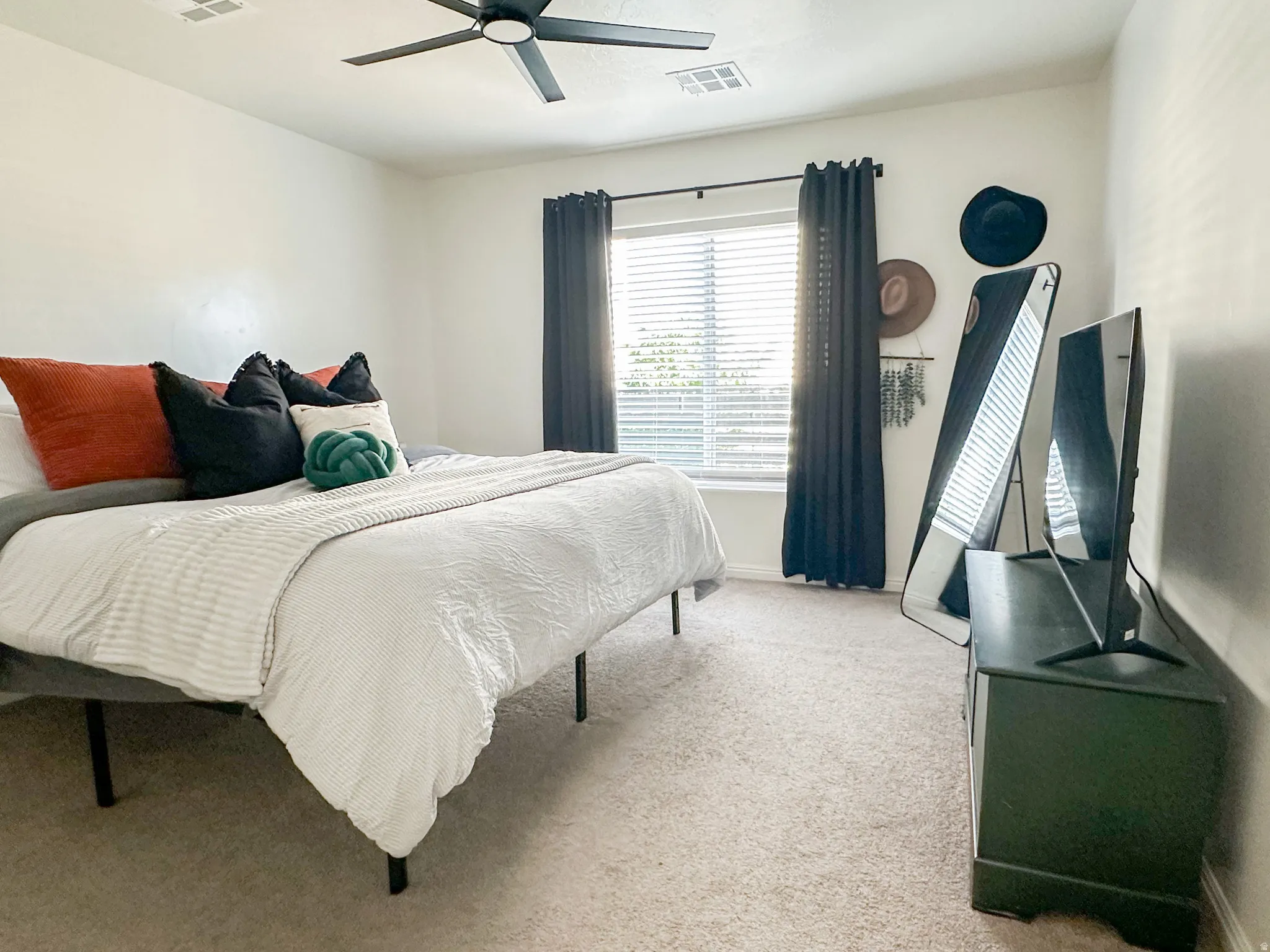Bedroom featuring light-colored walls, carpet flooring, and a large window with blinds and dark-colored curtains