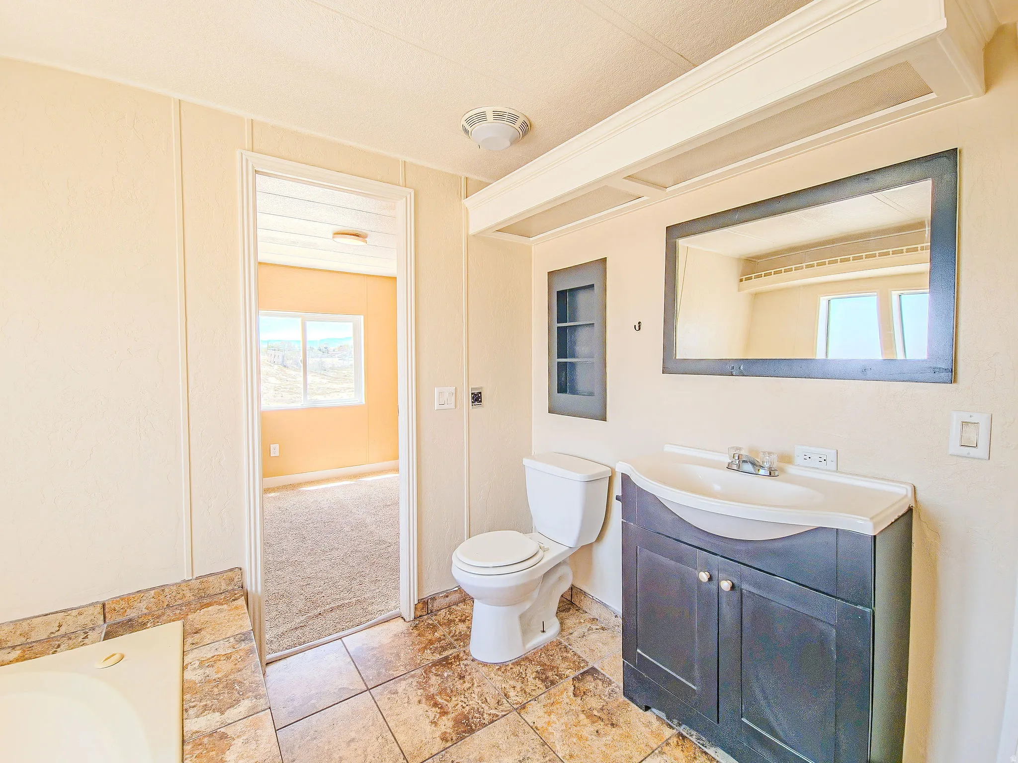 Bathroom featuring a dark wood-finish vanity with an integrated sink, framed mirror, built-in recessed shelving, and a jetted soaking tub with natural stone tile surround