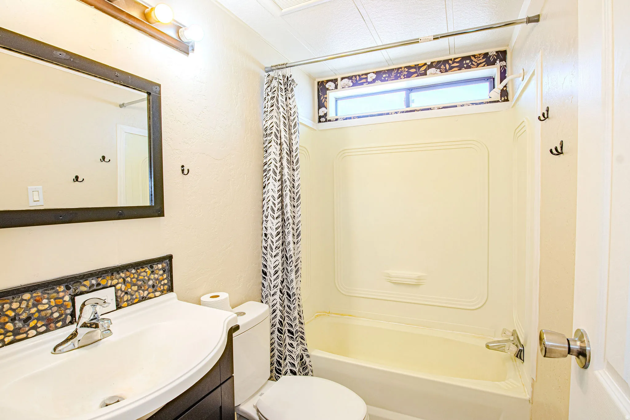Bathroom featuring a white bathtub with an integrated shower, a vanity with a mosaic pebble backsplash, a white ceramic sink, and a framed mirror