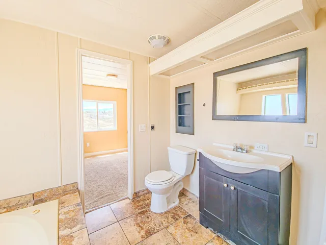 Bathroom featuring a dark wood-finish vanity with an integrated sink, framed mirror, built-in recessed shelving, and a jetted soaking tub with natural stone tile surround