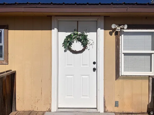 White four-panel entry door with black hardware, flanked by vertical siding and a single-pane window with blinds
