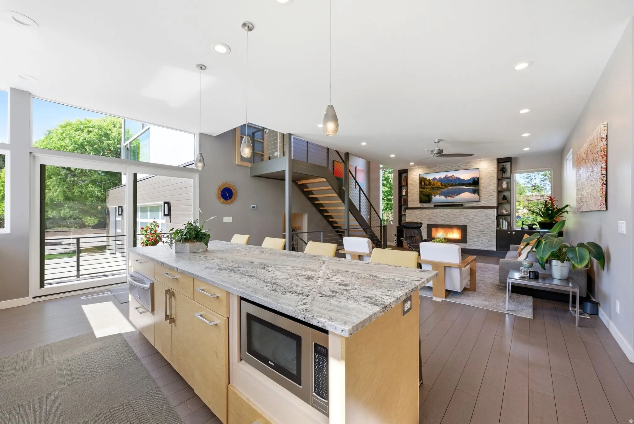 Spacious open-concept living area featuring a kitchen island with integrated microwave and dishwasher, extensive natural lighting from large windows and glass door, a modern fireplace with a tiled surround, and exposed metal stairs with wood treads