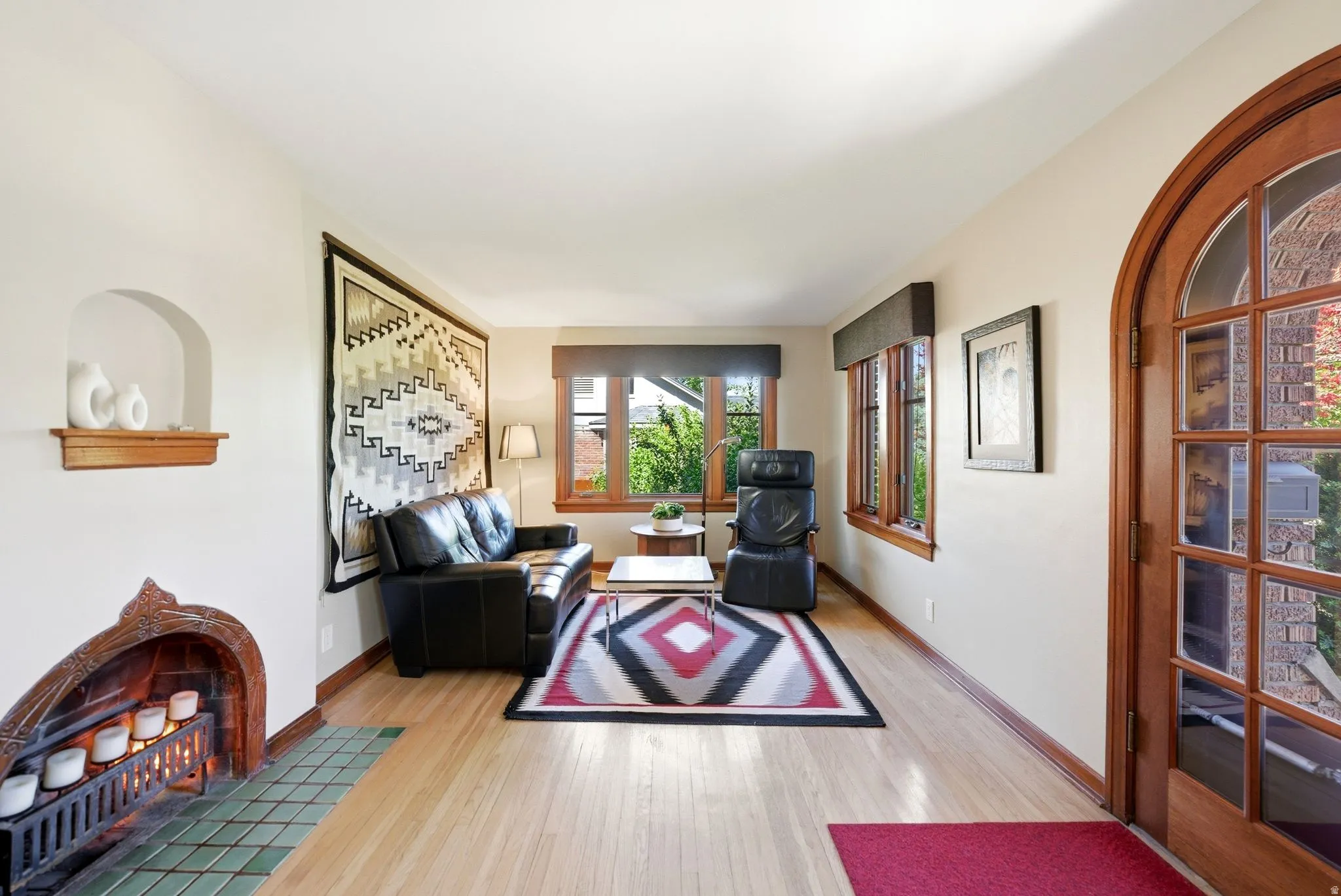 Arched wood-paneled entry door with glass inserts, wood-finish flooring, a built-in fireplace with decorative tile surround, an arched wall niche with a wood shelf, and multiple windows with wood trim