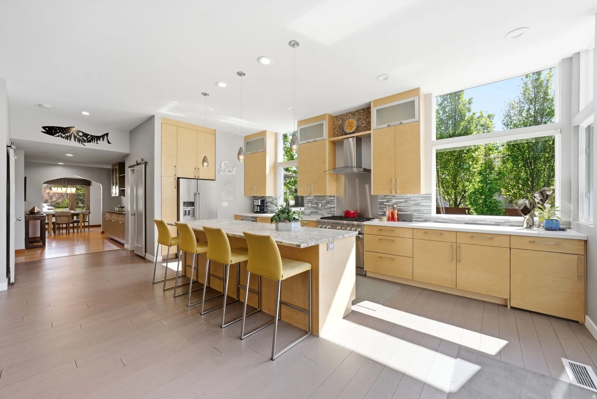 Modern kitchen featuring light wood cabinetry, a large center island with seating, a stainless steel range hood, and extensive natural light through large windows