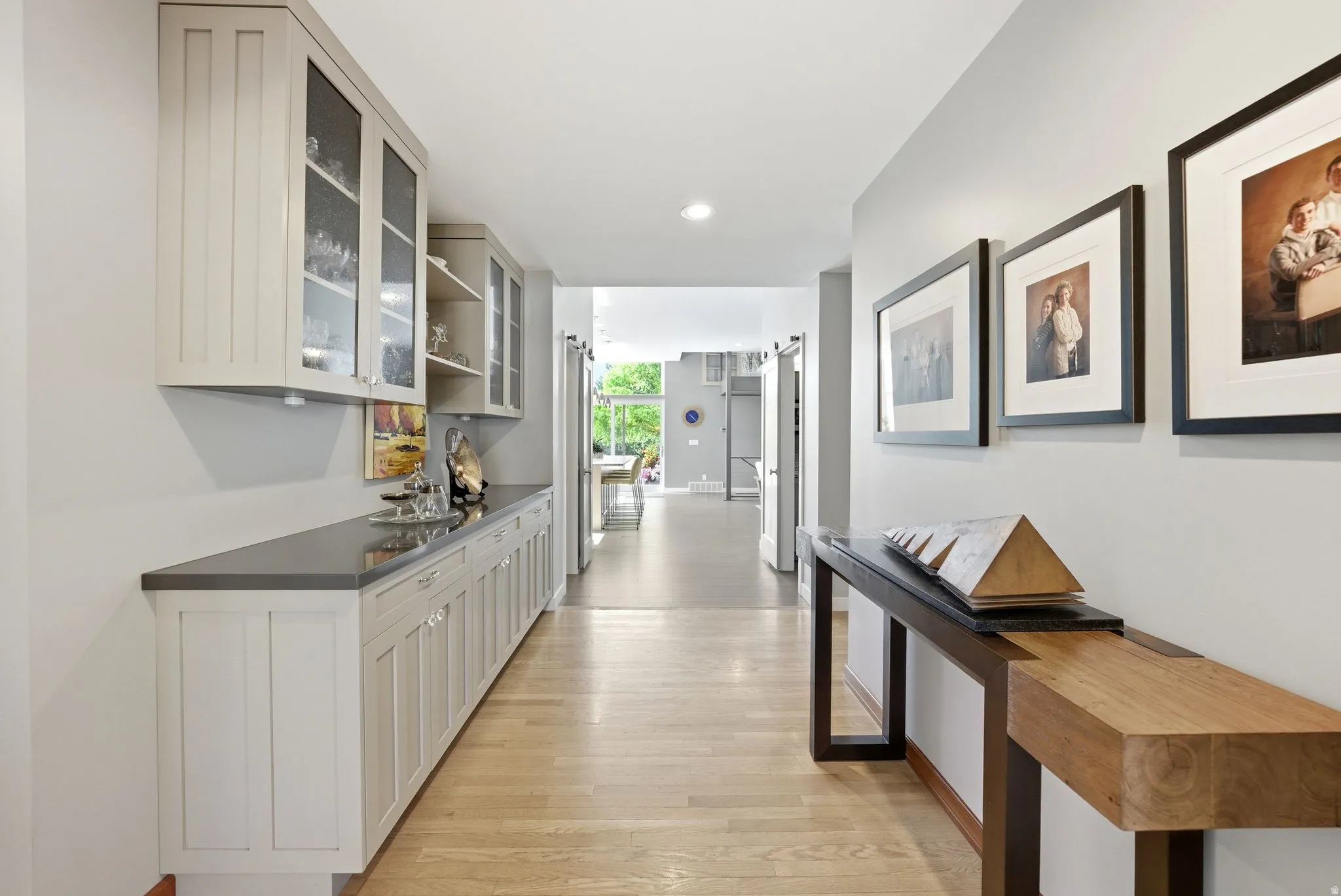 Hallway featuring wood-finish flooring, recessed lighting, built-in cabinetry with glass-front uppers, a dark countertop, and light gray wall paint