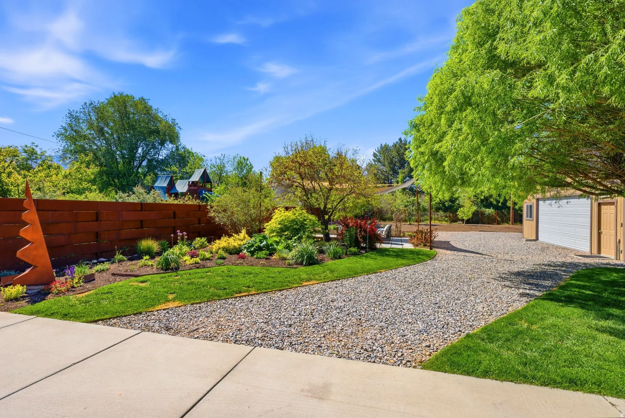 Gravel driveway with a tan detached garage, manicured lawn, and a retaining wall with integrated landscaping