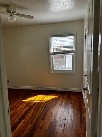 Room featuring wood-finish flooring, a window with blinds, a ceiling fan with light fixture, white trim, and a paneled door