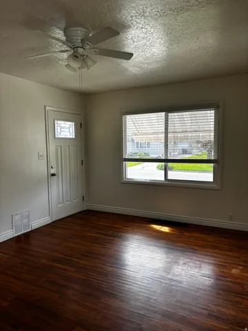 Room featuring rich wood-finish flooring, a white paneled entry door with decorative glass, a window with horizontal blinds, a white ceiling fan with integrated lighting, and white baseboards