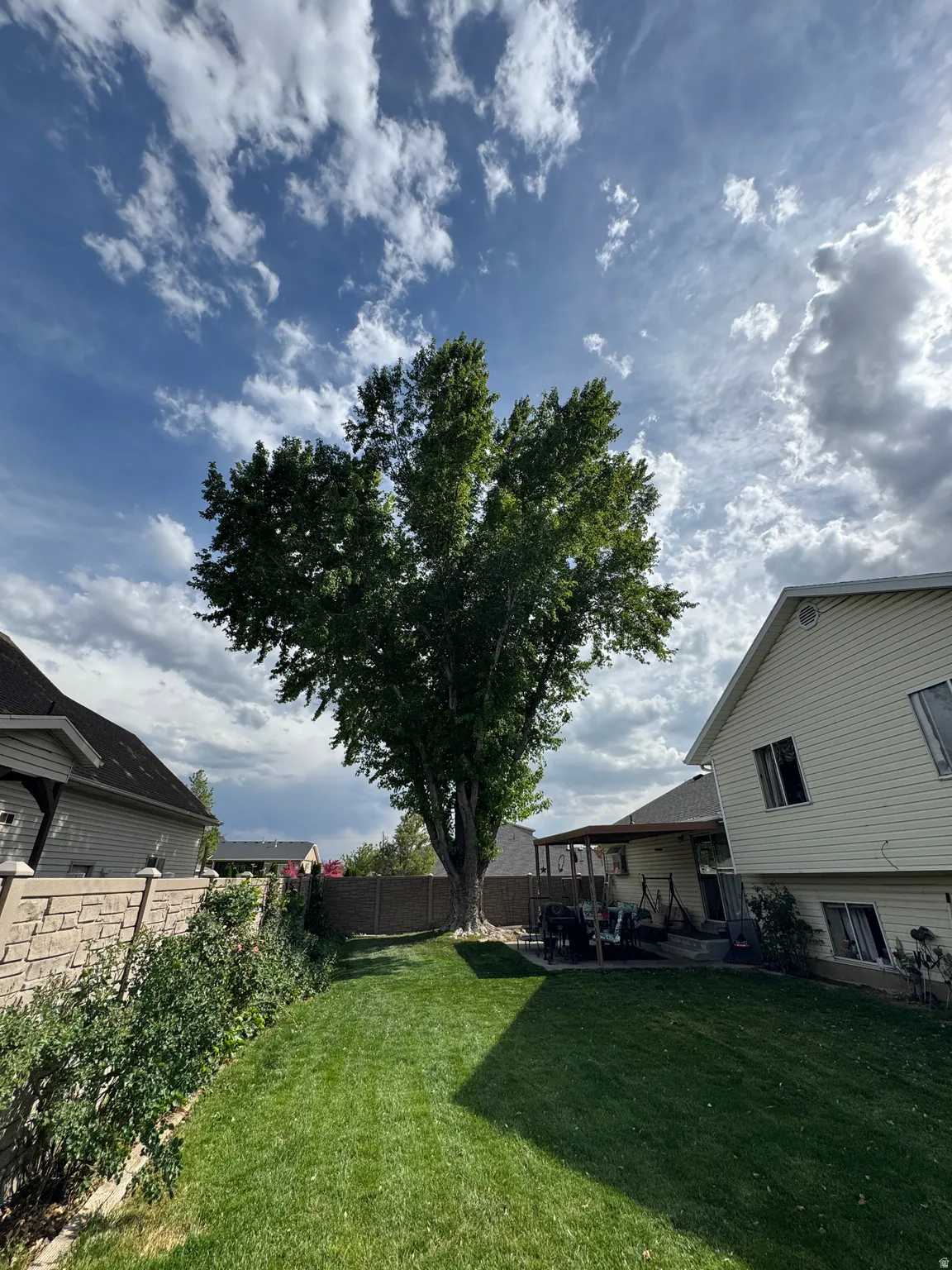 Spacious backyard featuring a substantial mature tree, a manicured lawn, a covered patio area, a wood fence, and a stone retaining wall