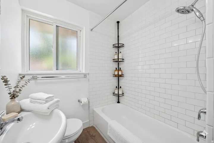 Bathroom featuring a white subway tile shower surround, a built-in tub, wood-finish flooring, a white vanity with a basin sink, and a sliding window
