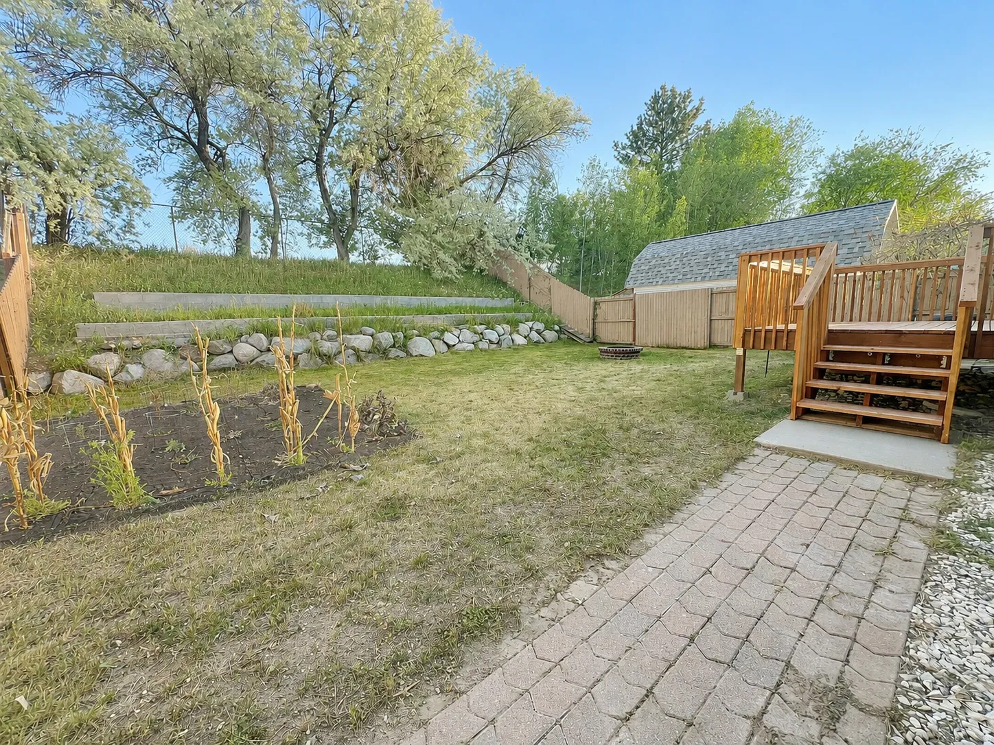 Backyard featuring a wood deck with stairs, a paved pathway, a garden bed, a rock retaining wall, and a wood fence