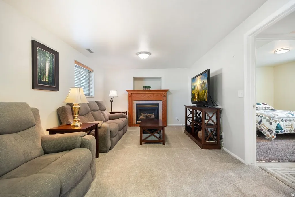 Living area featuring a fireplace with a wood mantle, light-colored carpeting, a window with blinds, and an open doorway to an additional room