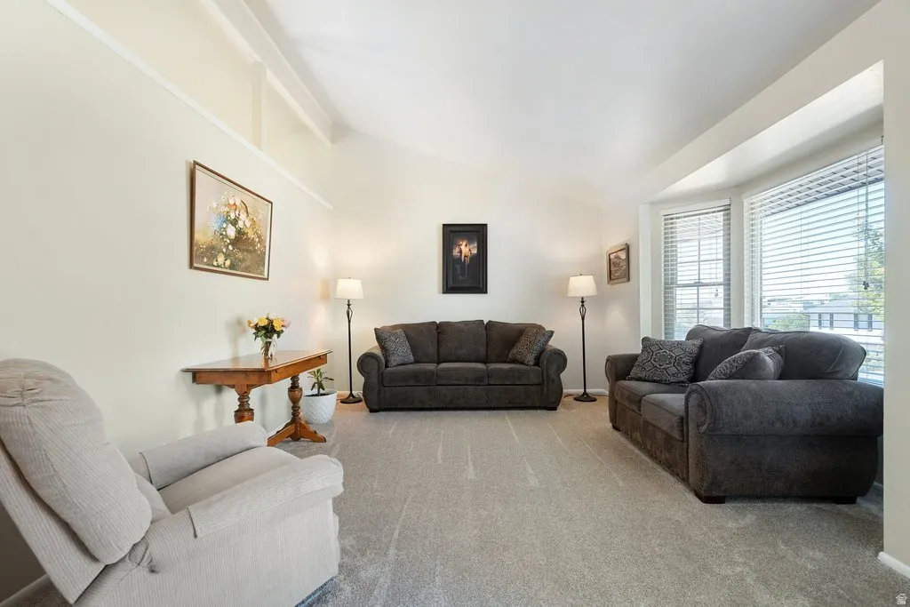 Spacious living area featuring a vaulted ceiling with exposed beams, light-colored walls, and neutral carpeting