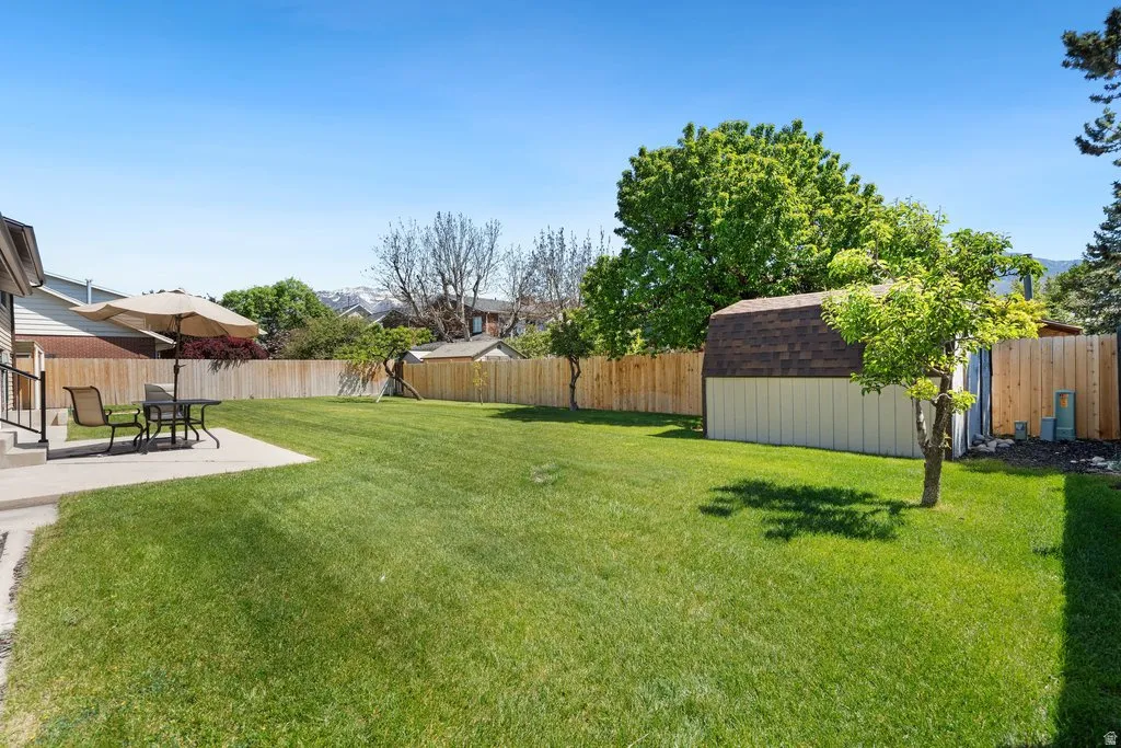 Spacious lawn area with a concrete patio, a privacy fence, and a storage shed with a shingle roof