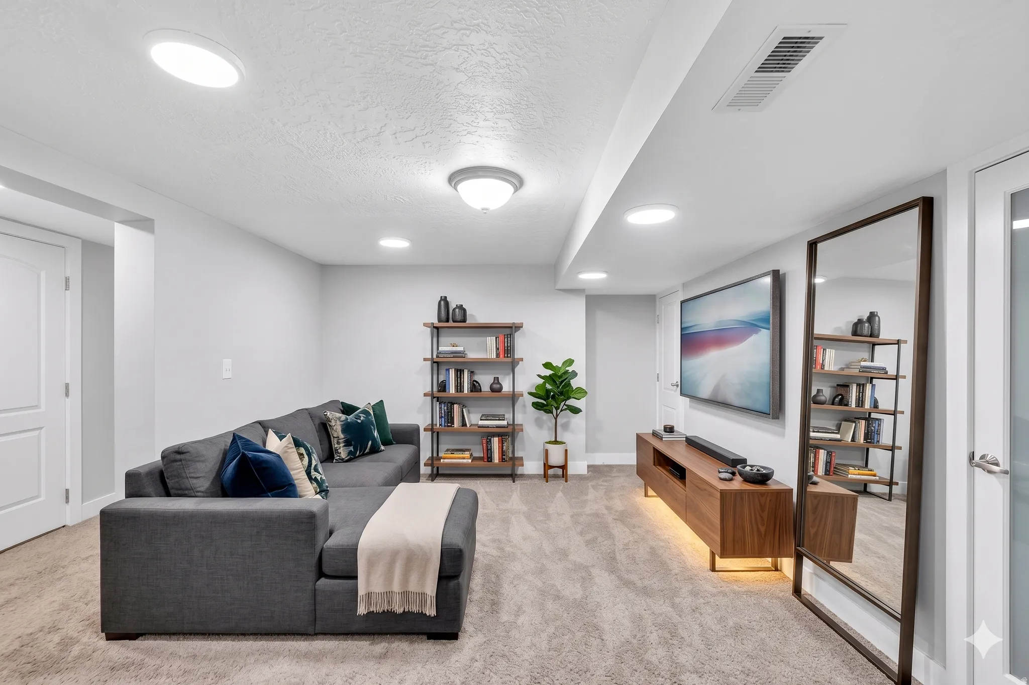 Recessed lighting illuminates this flexible living area with neutral carpeting, white walls, and a modern wood media console featuring integrated lighting