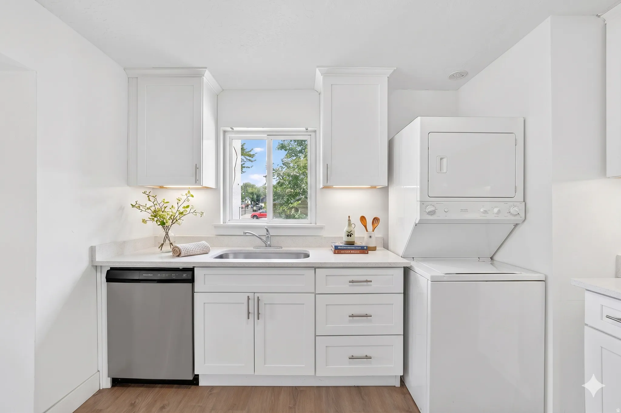 Updated kitchen area featuring white shaker cabinetry, light-toned countertops, a stainless steel dishwasher, a single-basin sink with a chrome faucet, and wood-finish flooring