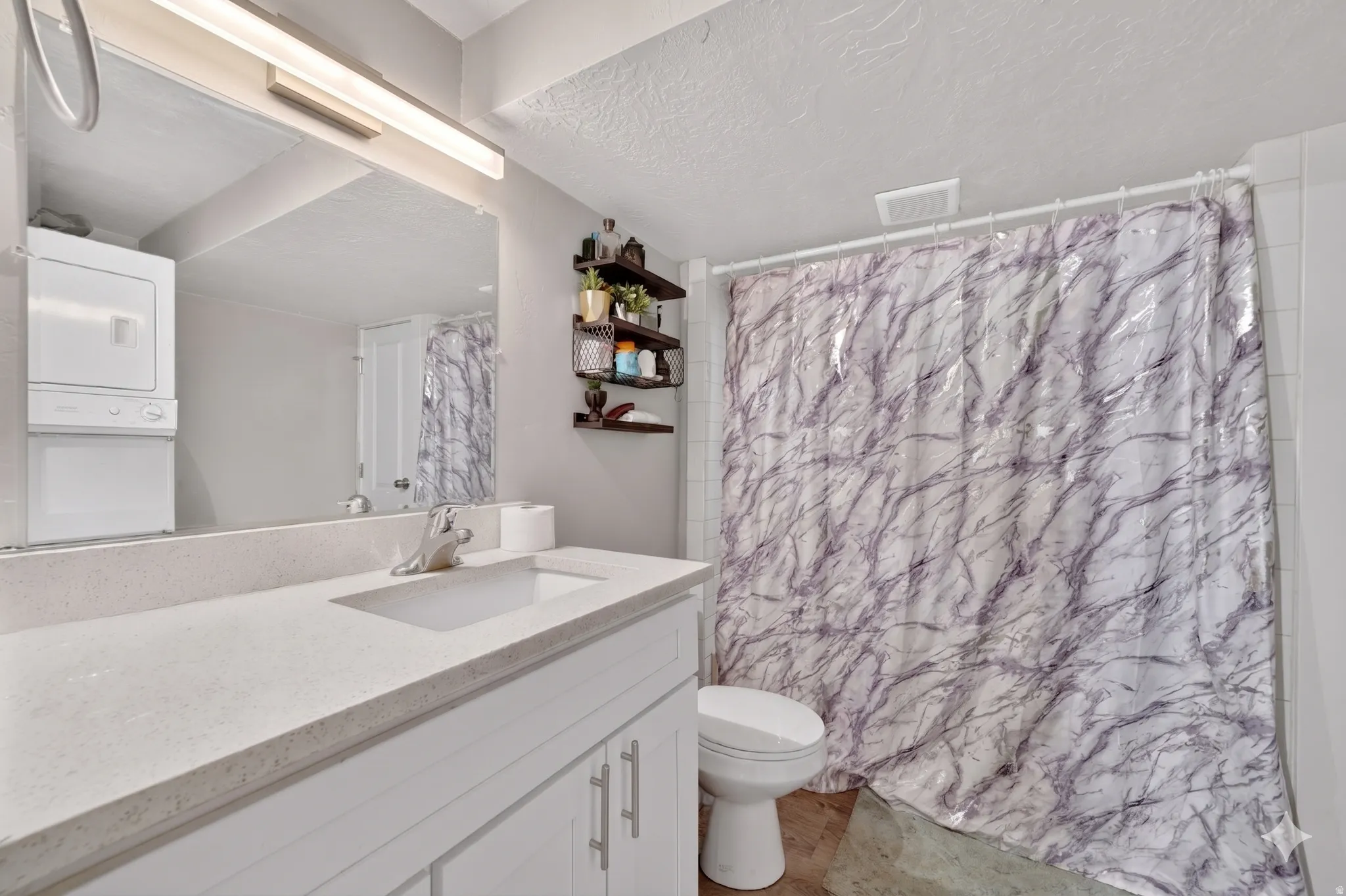 Bathroom featuring a single vanity with a light-toned countertop, an integrated sink, a large wall-mounted mirror, and a stacked washer and dryer unit