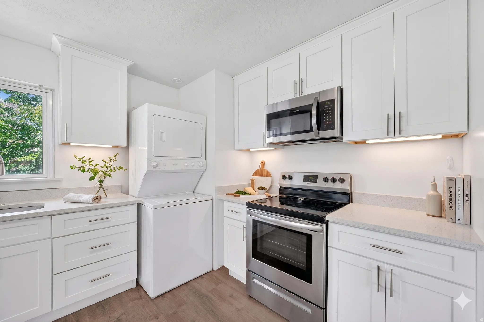 Bright kitchen featuring white shaker cabinetry, stainless steel appliances, stacked laundry machines, light-toned countertops, and wood-finish flooring