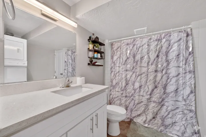 Bathroom featuring a single vanity with a light-toned countertop, an integrated sink, a large wall-mounted mirror, and a stacked washer and dryer unit