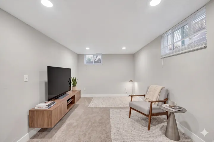 Recessed ceiling lighting illuminates this versatile space featuring light gray walls, plush carpet, and a built-in wood-finish media console