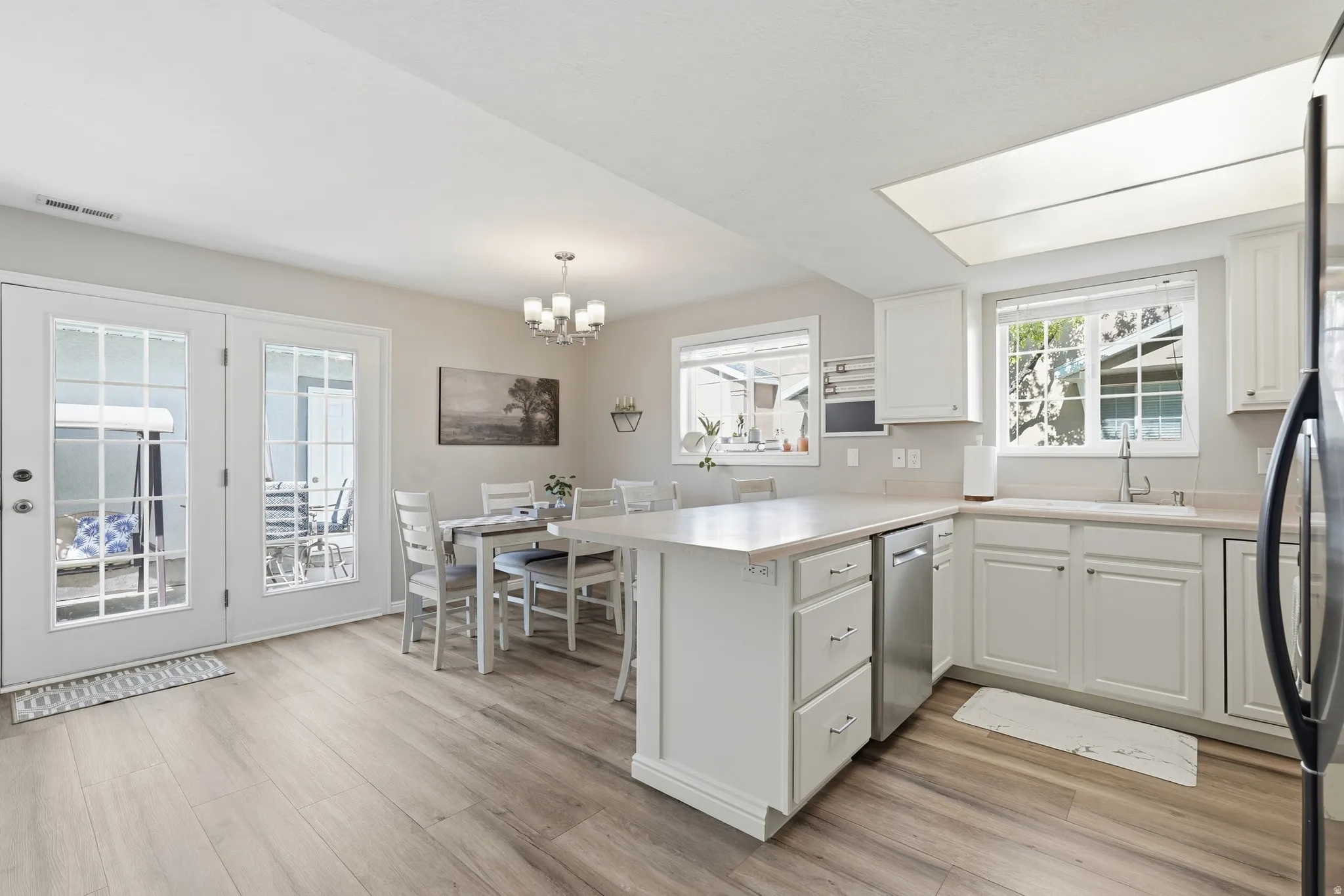 Bright kitchen and dining area featuring light wood-finish flooring, white cabinetry, a built-in stainless steel dishwasher, and a large window above the sink
