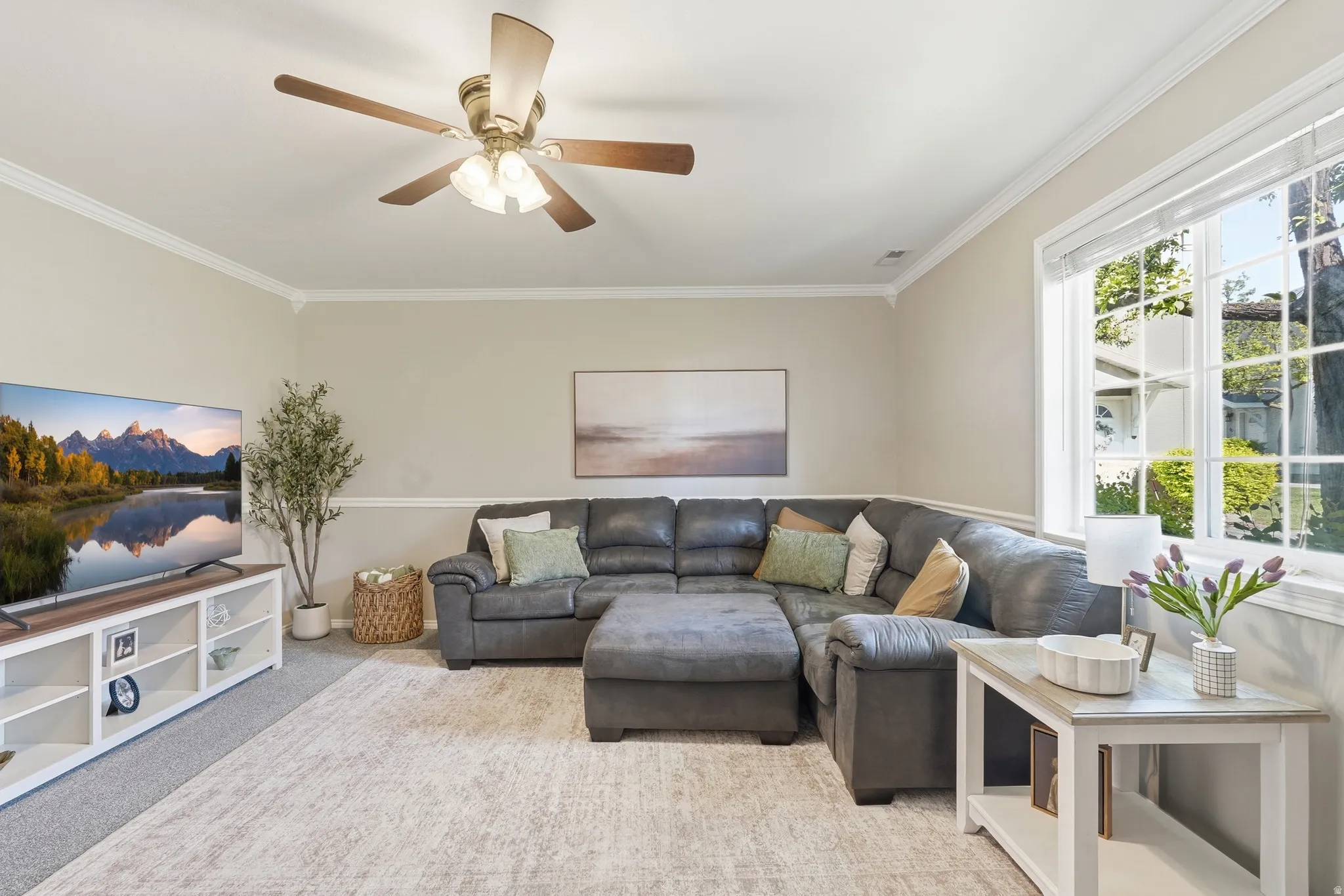 Spacious living area featuring light-toned walls, crown molding, a ceiling fan with integrated lighting, and a large multi-pane window