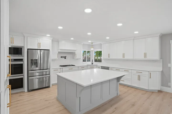 Spacious kitchen featuring an expansive central island with a light-toned countertop, ample white cabinetry with gold-tone hardware, stainless steel appliances, and light wood-finish flooring