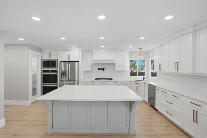Spacious kitchen featuring a large central island with a light-toned countertop, white shaker cabinetry with gold-toned hardware, stainless steel appliances, and light wood-finish flooring