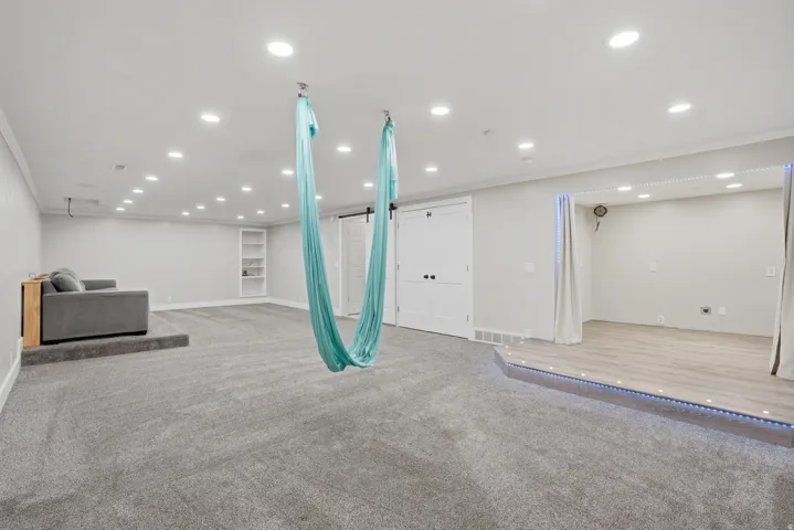 Spacious lower-level area with recessed lighting, built-in shelving, carpet flooring, and a raised platform with wood-finish flooring and integrated lighting