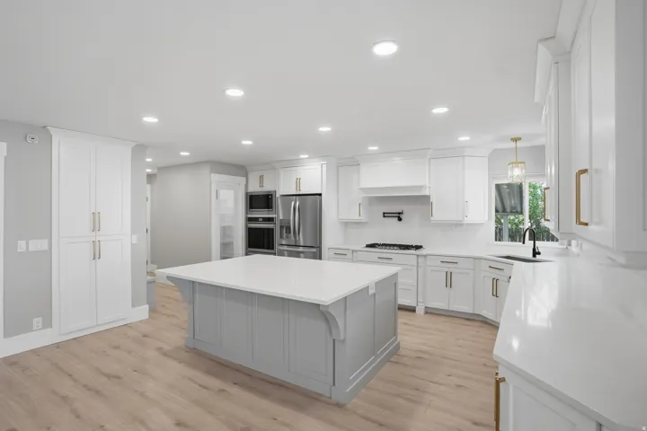 Kitchen featuring a light gray island with a white countertop, white perimeter cabinetry with gold-tone hardware, stainless steel appliances, and wood-finish flooring
