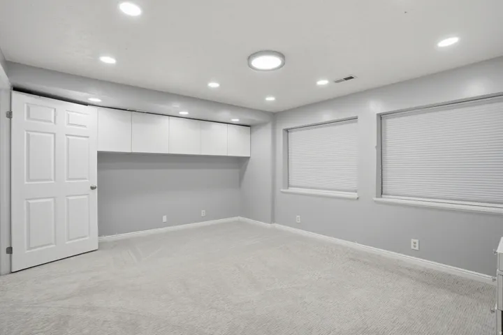 Spacious room featuring light gray carpeting, recessed lighting, two windows with blinds, and white built-in cabinetry with under-cabinet lighting