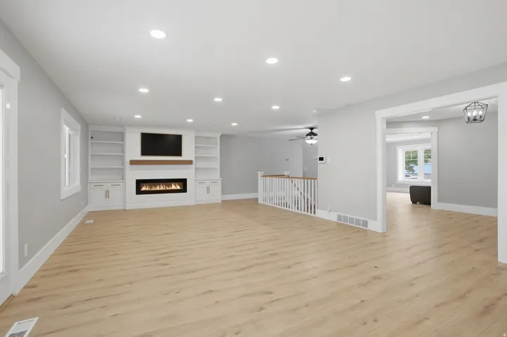 Spacious living area featuring light wood-finish flooring, a contemporary fireplace with a wood mantle, built-in shelving, recessed lighting, and a white staircase with a contrasting wood handrail
