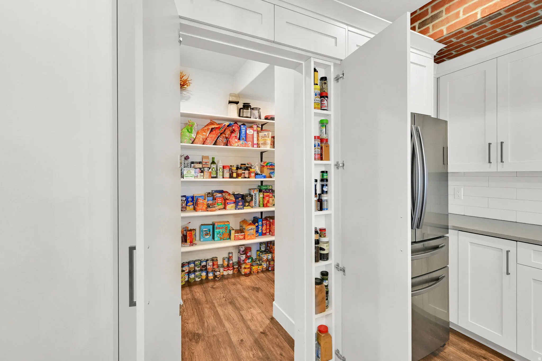 Walk-in pantry with extensive shelving, wood-finish flooring, and a built-in spice rack, complemented by white shaker-style cabinetry, stainless steel refrigerator, and a subway tile backsplash